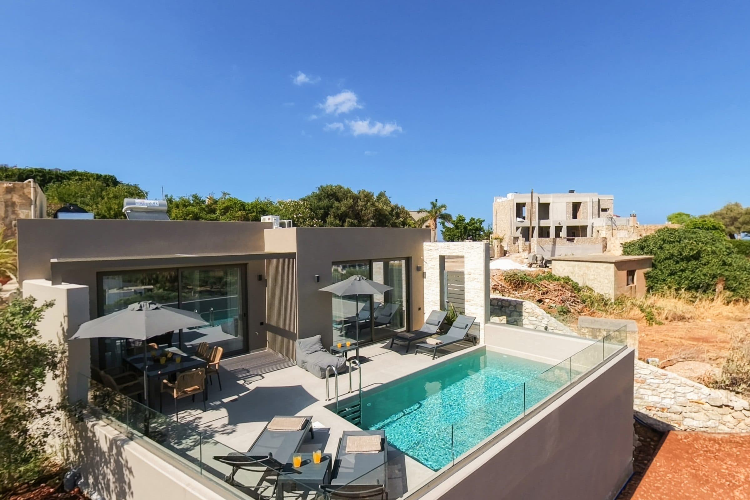 Elevated view of the villa with its shaded outdoor dining table, sun loungers and rectangular pool enclosed by a glass balustrade