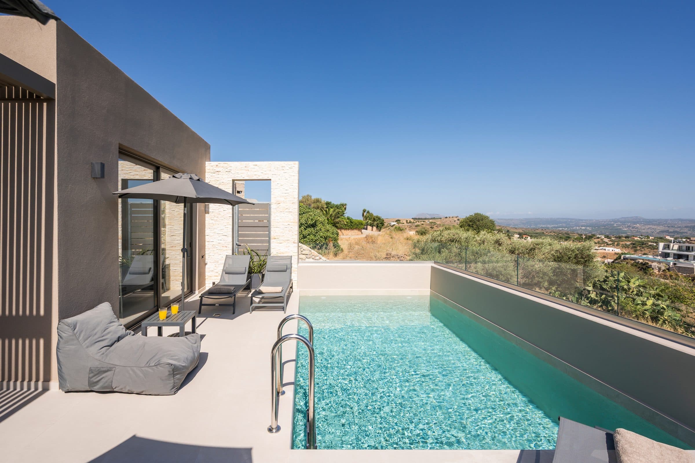 The pool seen along its length, with a grey bean bag, sun loungers and a parasol on the terrace and open countryside beyond
