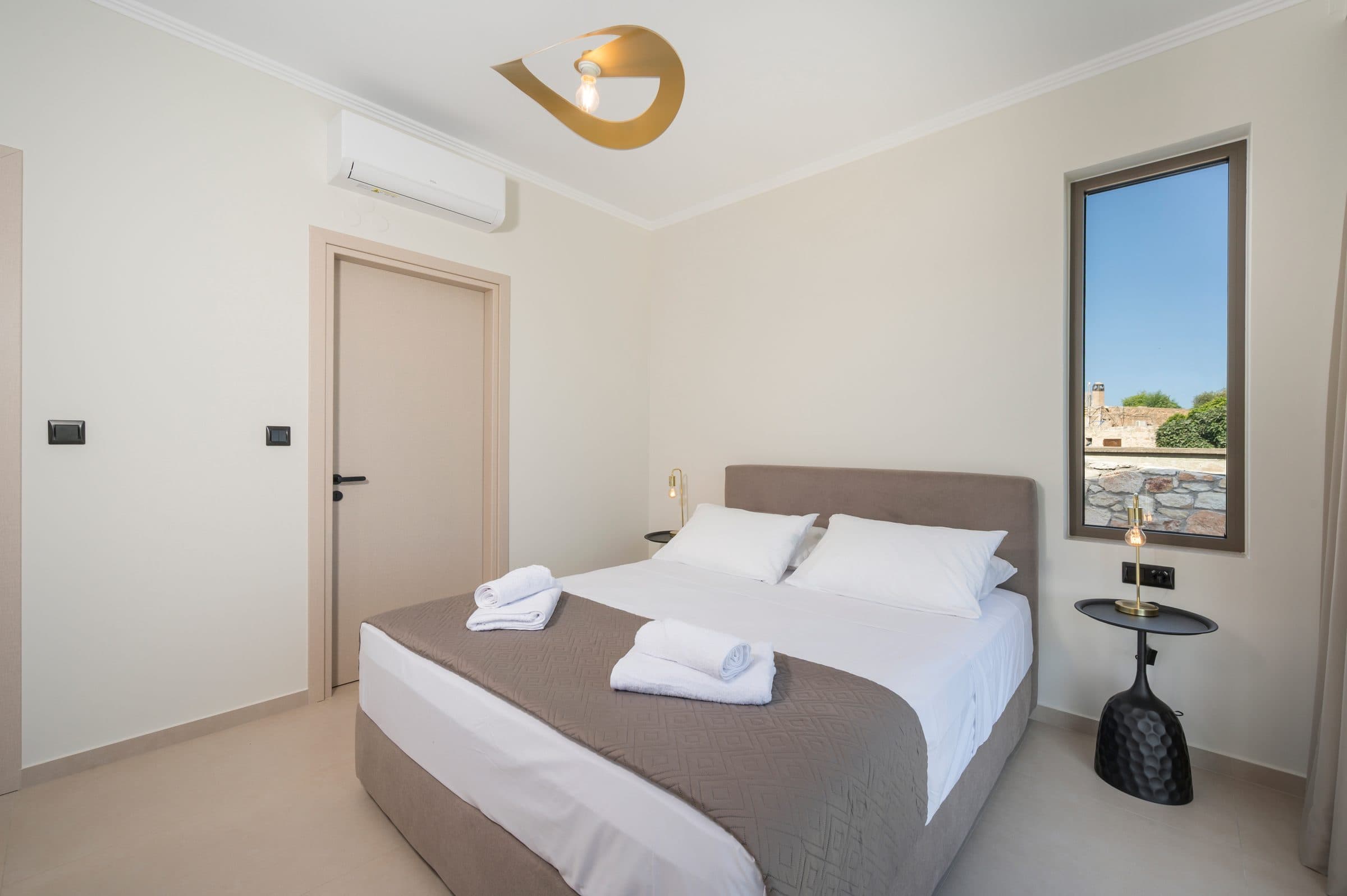 The second bedroom with a queen bed, air conditioning and a tall narrow window framing the stone courtyard wall