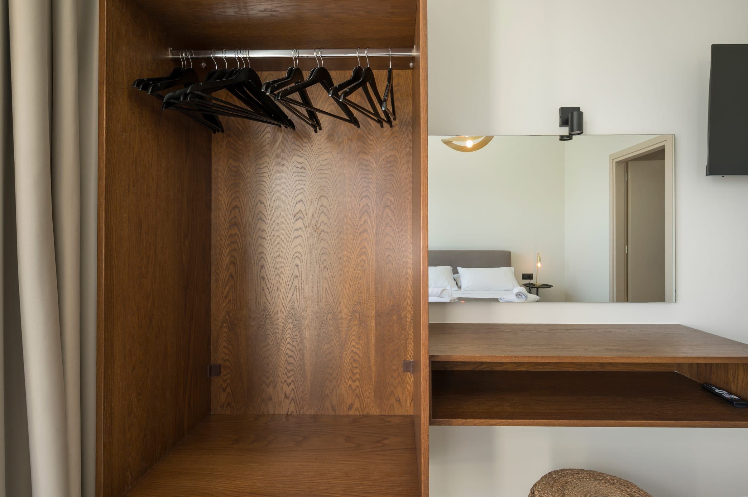 The open oak wardrobe with hangers, beside the bedroom mirror reflecting the bed