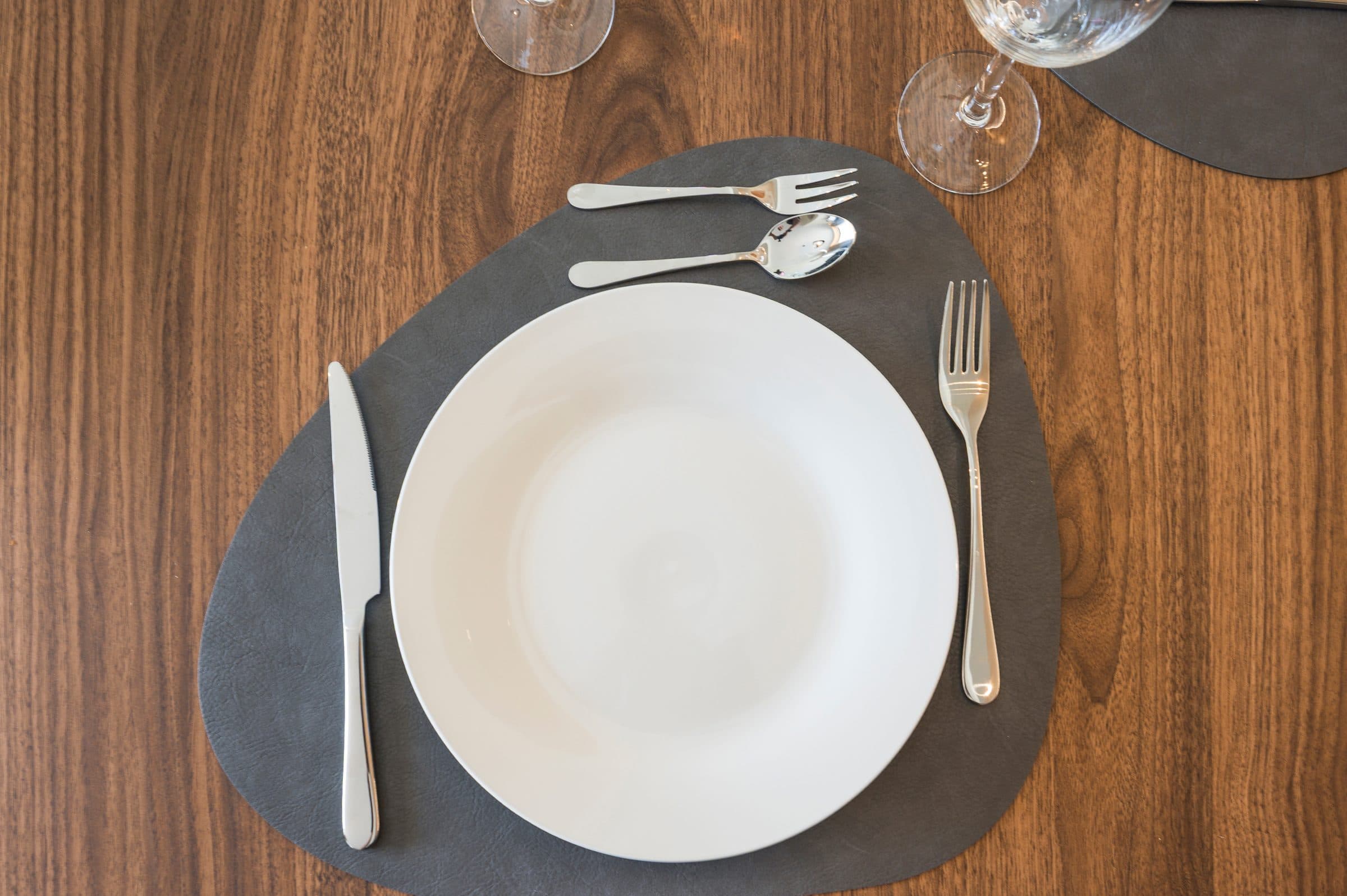 A place setting of white china and polished cutlery on a leather mat over the walnut dining table