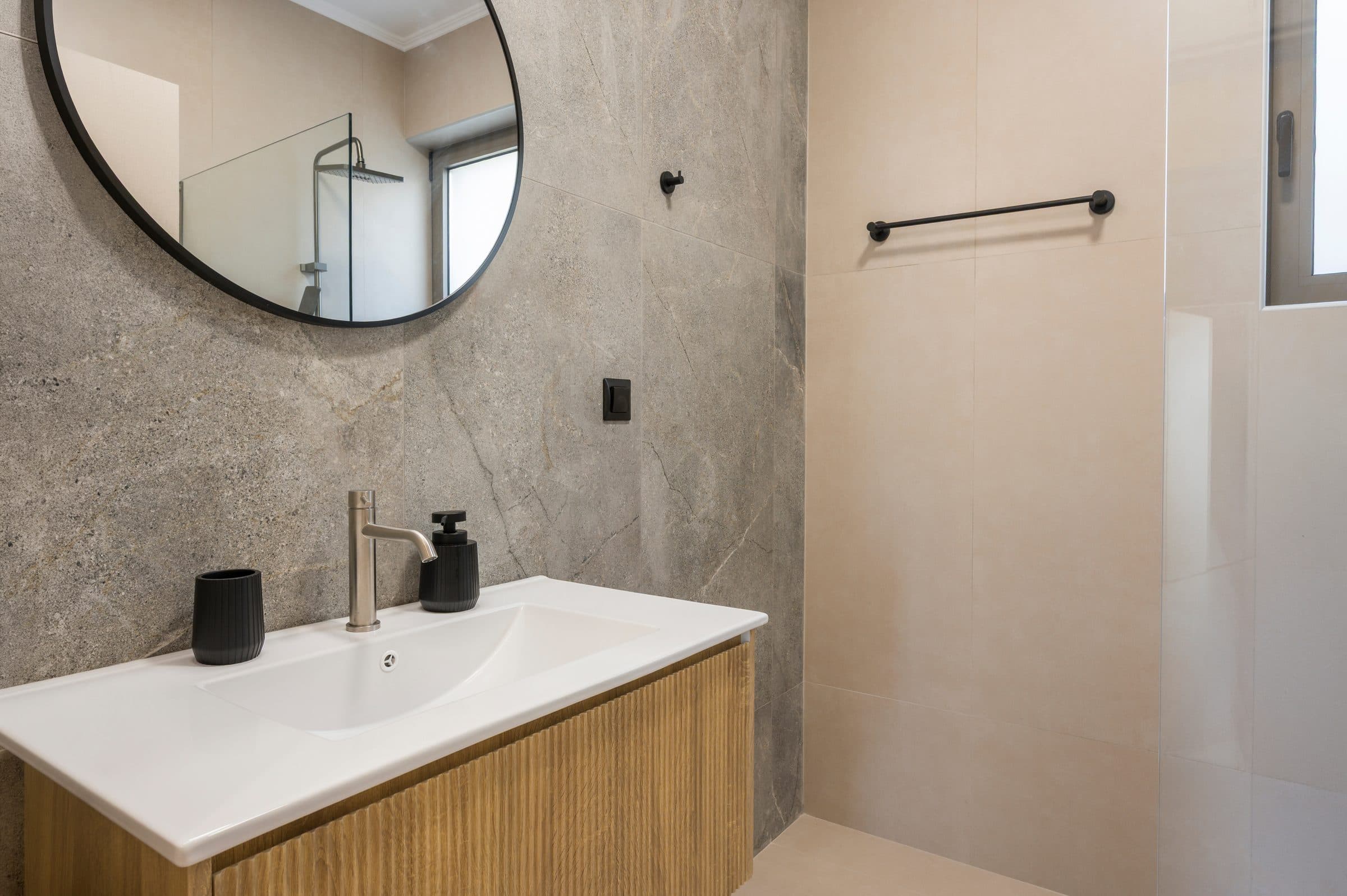 A bathroom in stone-effect tile with a black round mirror, oak vanity unit and glass walk-in shower
