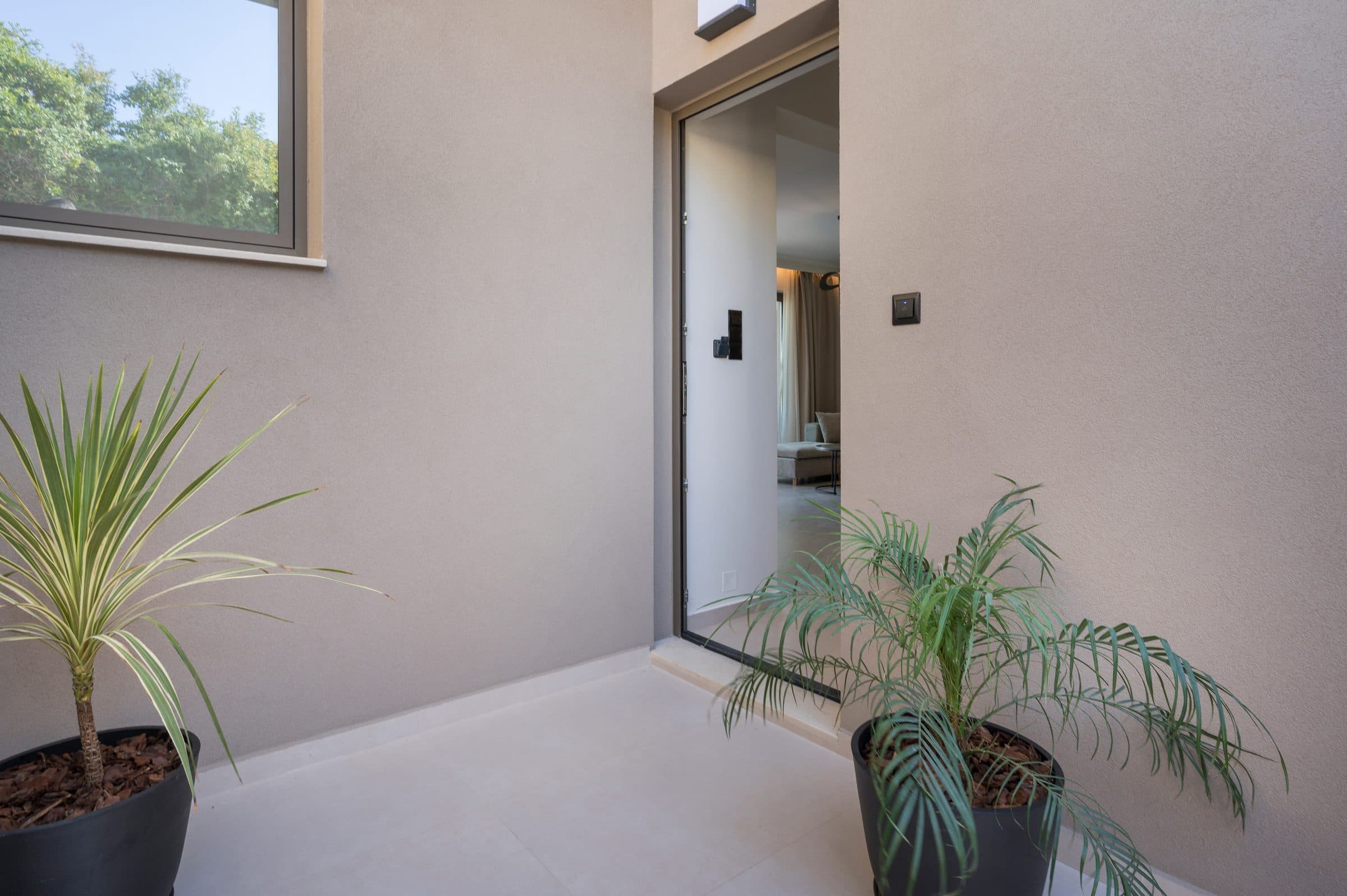 The villa entrance: a tall dark-framed door flanked by potted palms against smooth sand-coloured plaster