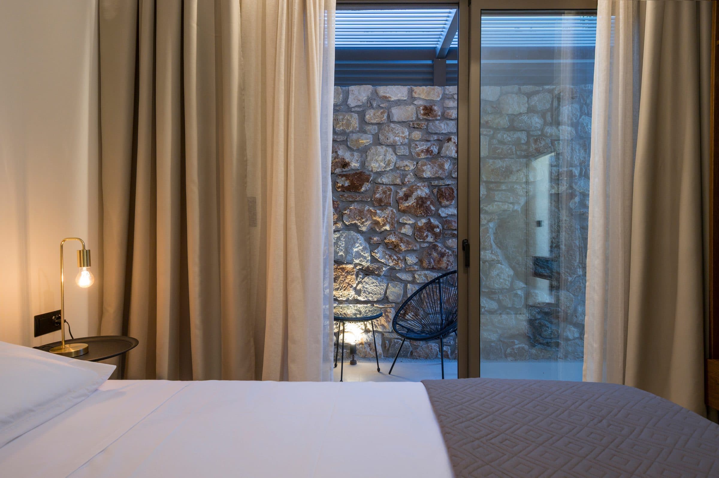 A bedroom at dusk, a brass bedside lamp lit and the sliding door framing the stone courtyard beyond