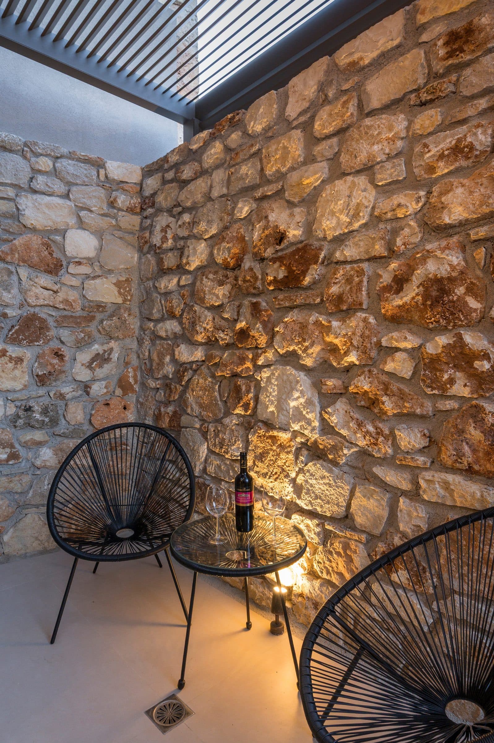 Two black wire chairs and a bottle of red wine on a glass table against the warmly lit stone wall of the courtyard