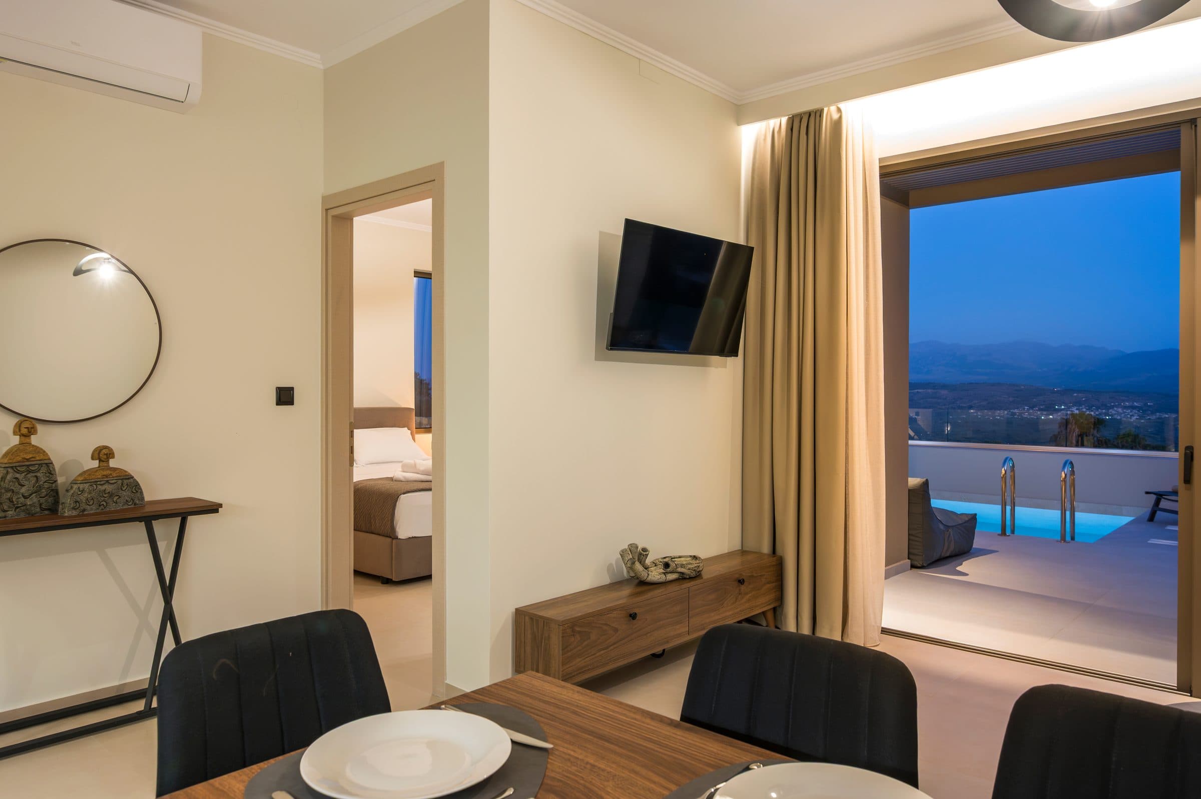 The interior after sunset, looking past the dining table and open bedroom door to the lit pool terrace