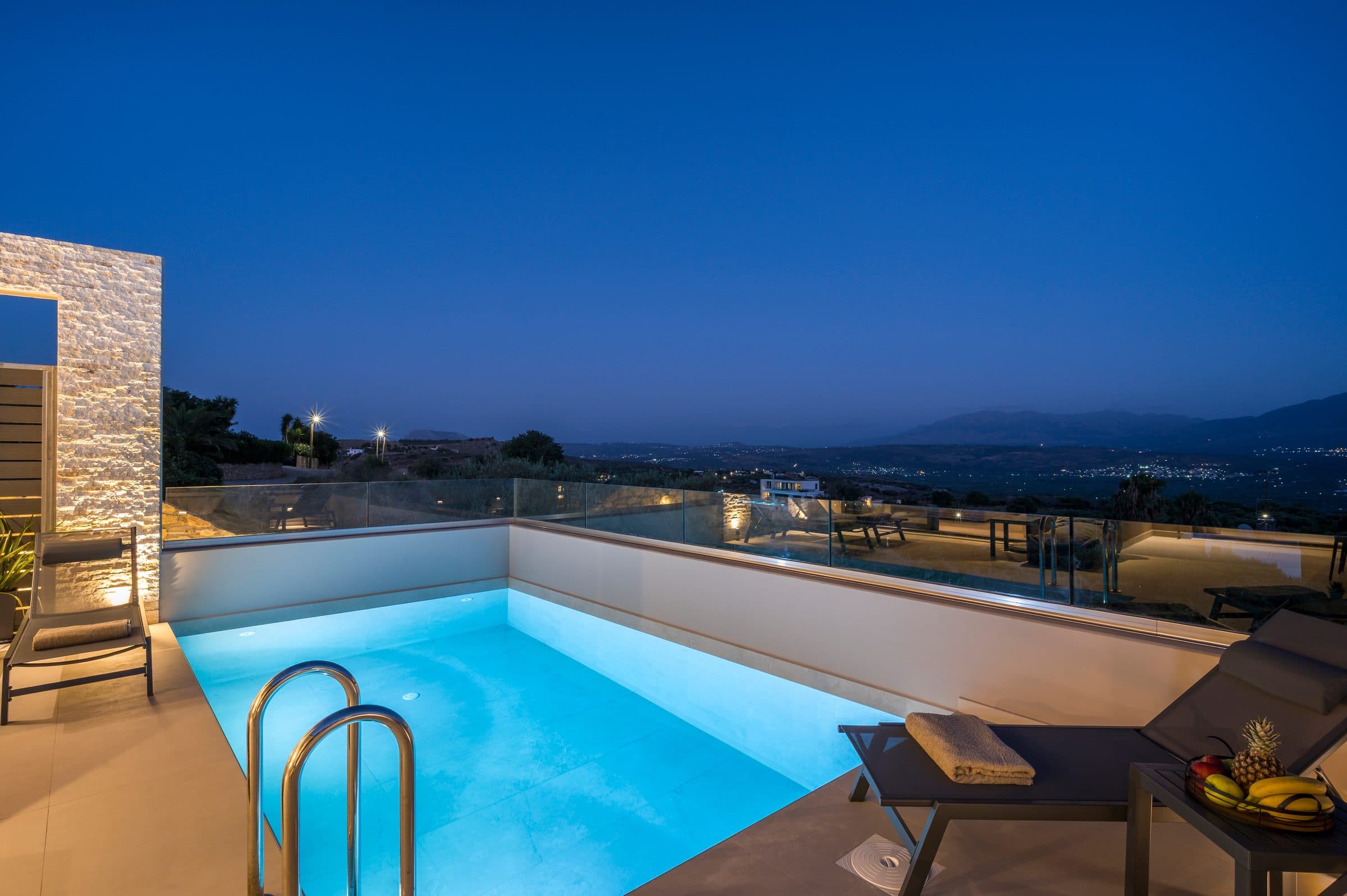 The illuminated pool and terrace at dusk, the glass balustrade opening onto the lights of the valley below