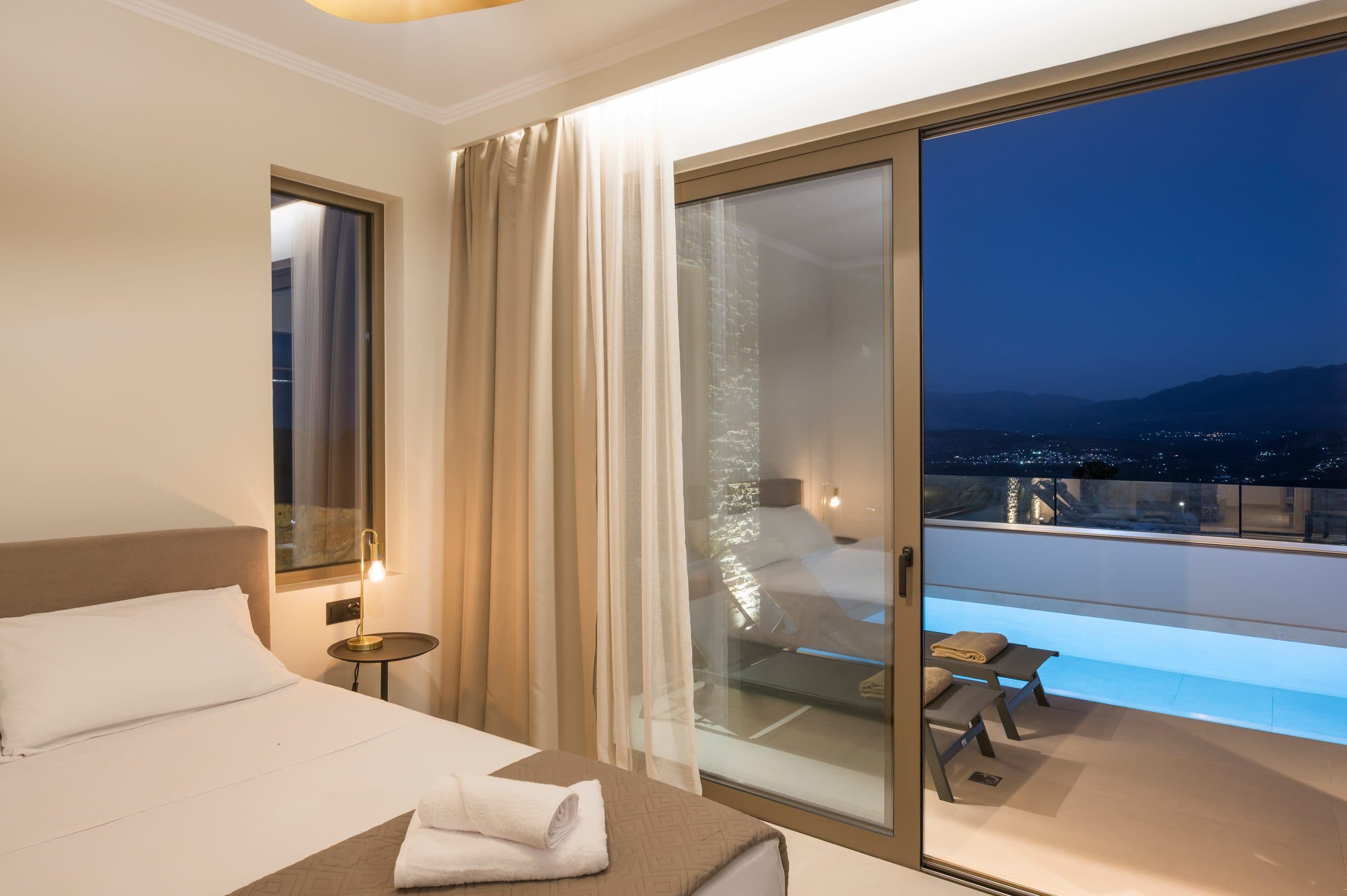 A bedroom at night, its sliding doors open onto the glowing pool and the distant lights of the valley