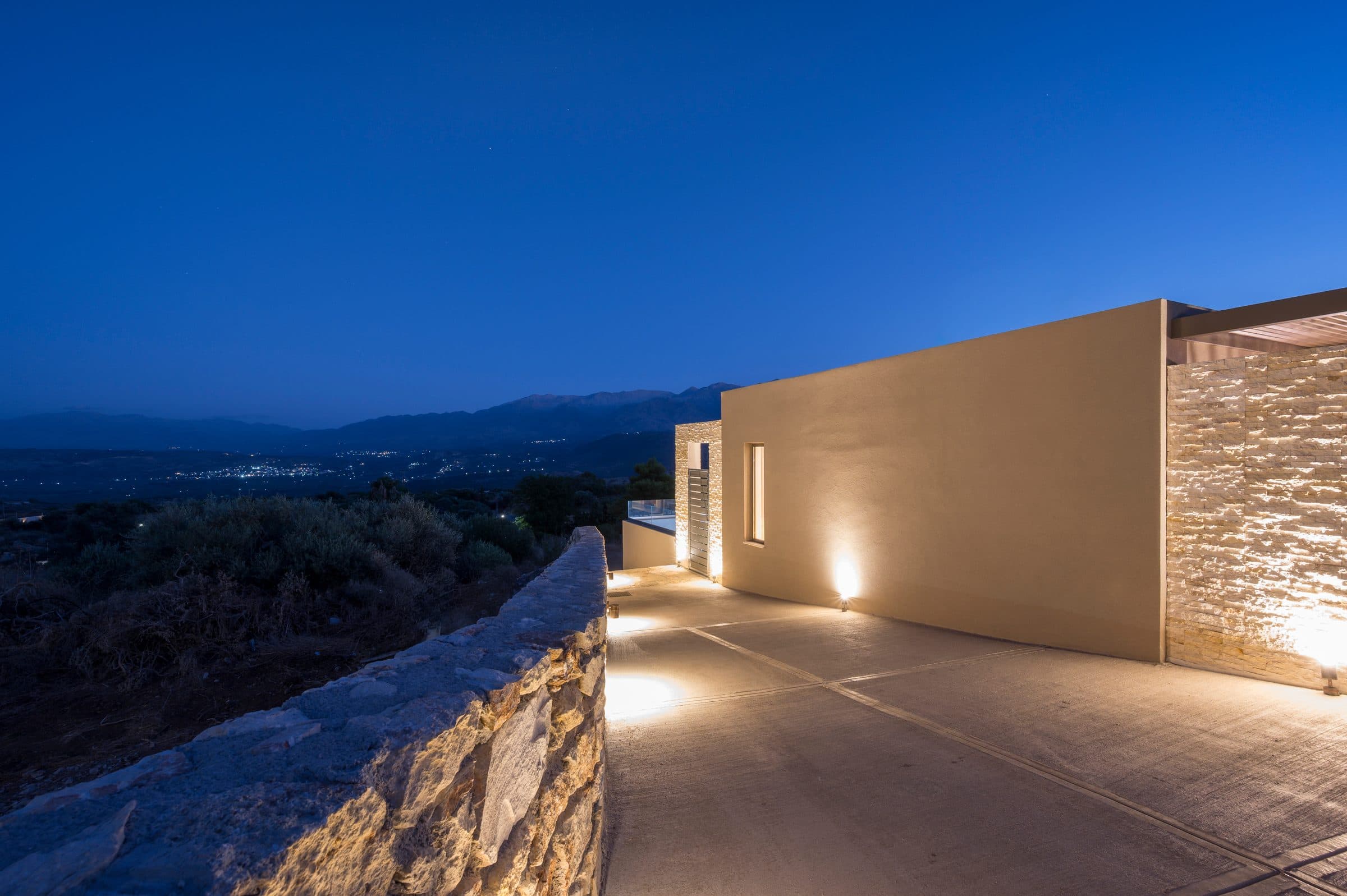 The approach to the villa at nightfall, the stone wall and plaster façade washed with warm light beneath a deep blue sky