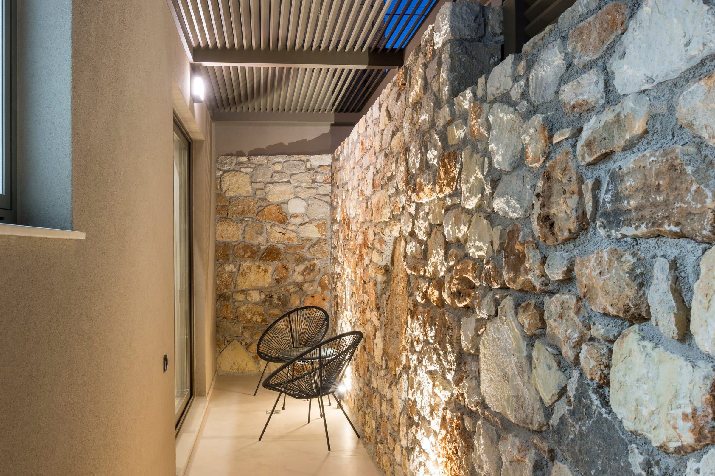 The traditional Cretan dry-stone wall of the side courtyard, uplit at dusk beneath the slatted pergola