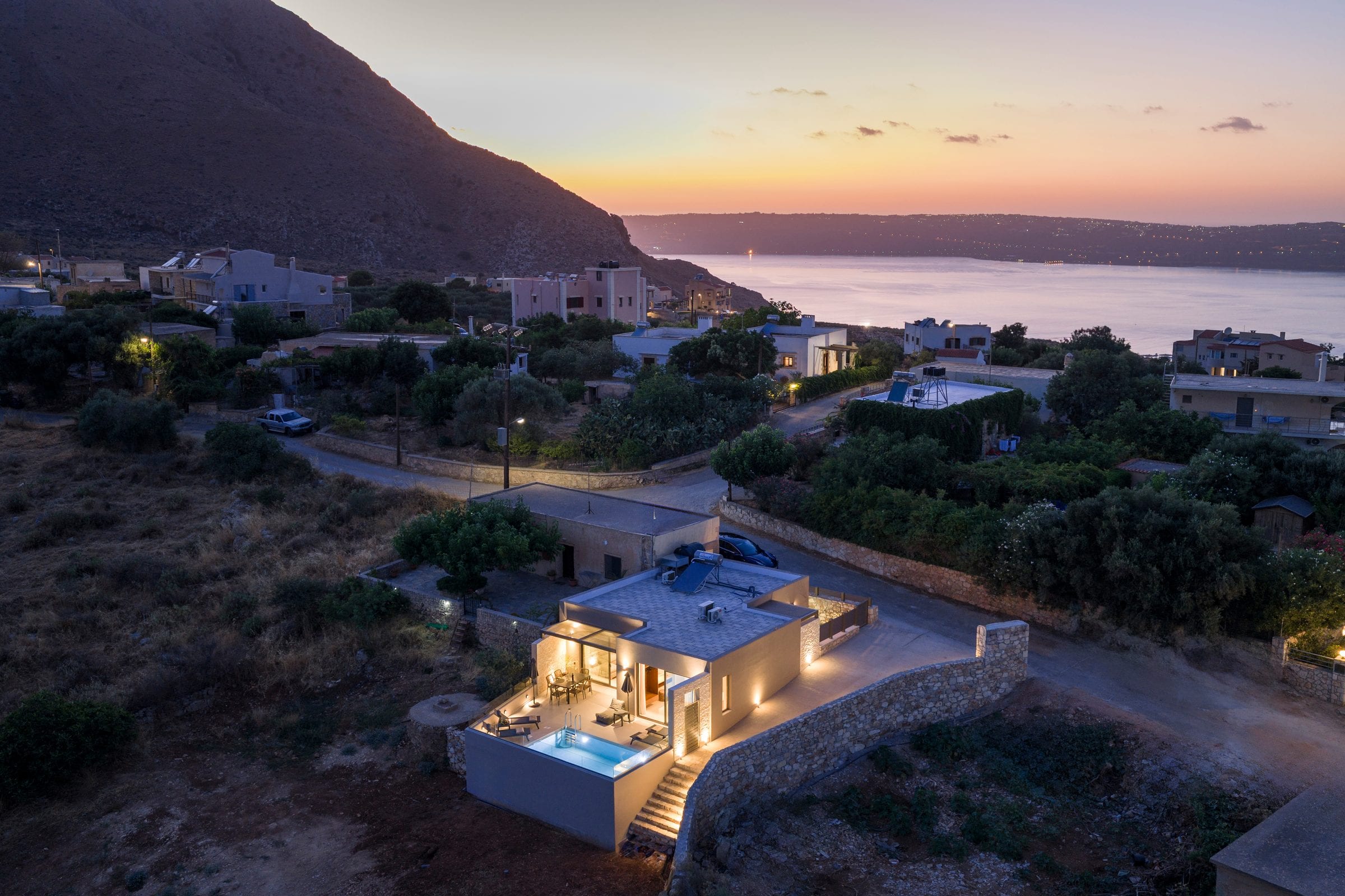 Aerial view at sunset of Casa Aura lit up on the hillside of Megala Chorafia, its pool glowing turquoise, with the village and Souda Bay behind it