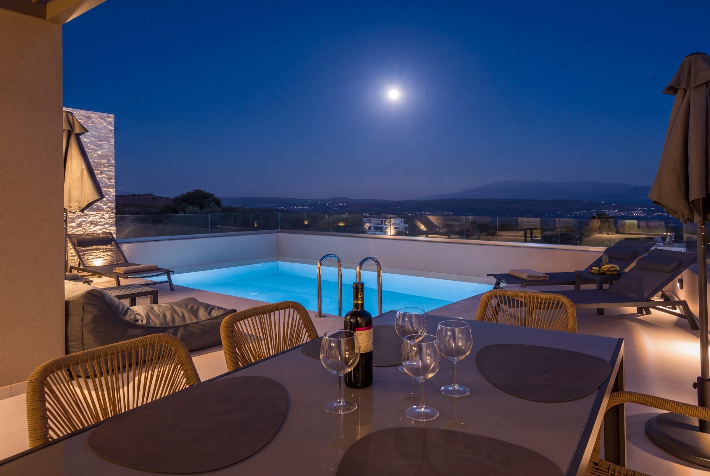 The terrace dining table set with wine glasses beside the illuminated pool under a full moon